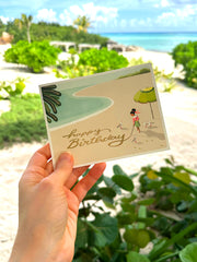 Woman writing on sand in a tropical beach birthday card by JooJoo Paper